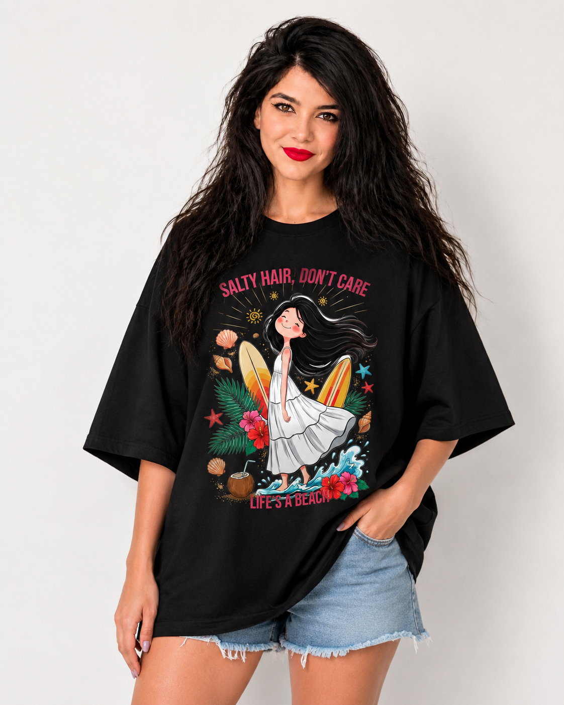 A woman modeling an oversized black graphic t-shirt with a tropical beach illustration and the text 'SALTY HAIR, DON'T CARE' and 'LIVE LIKE A LOCAL'. She is wearing light-wash denim shorts against a plain light gray background.
