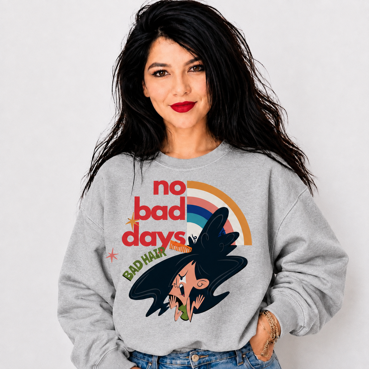A woman with long black hair wearing a light gray sweatshirt with a retro graphic that reads 'no bad days' and 'BAD HAIR' with a rainbow and cartoon witch illustration, paired with blue jeans against a light gray background.