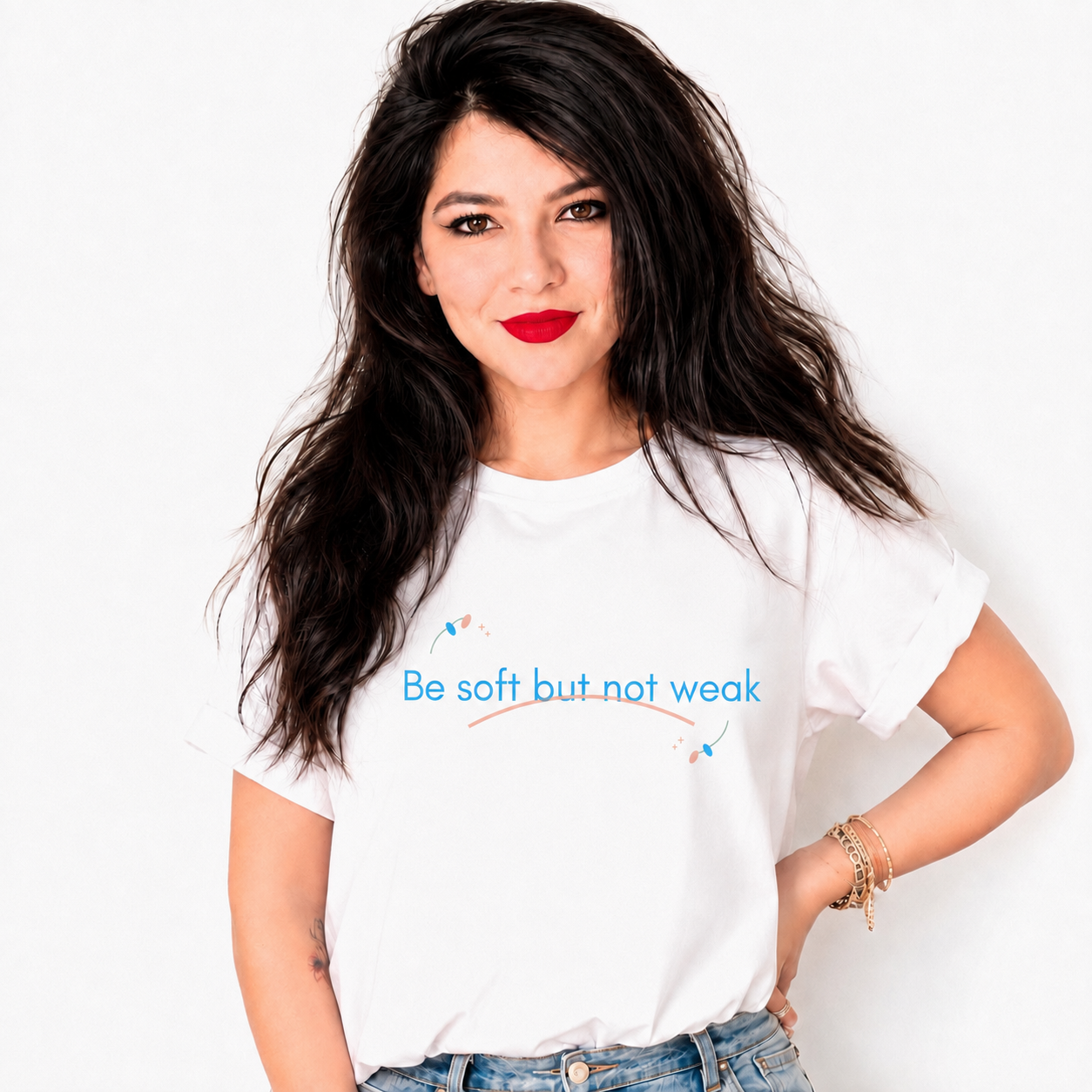 A woman with long dark hair and red lipstick wearing a white graphic t-shirt with the text 'Be soft but not weak' in blue, paired with blue jeans, against a plain white background.