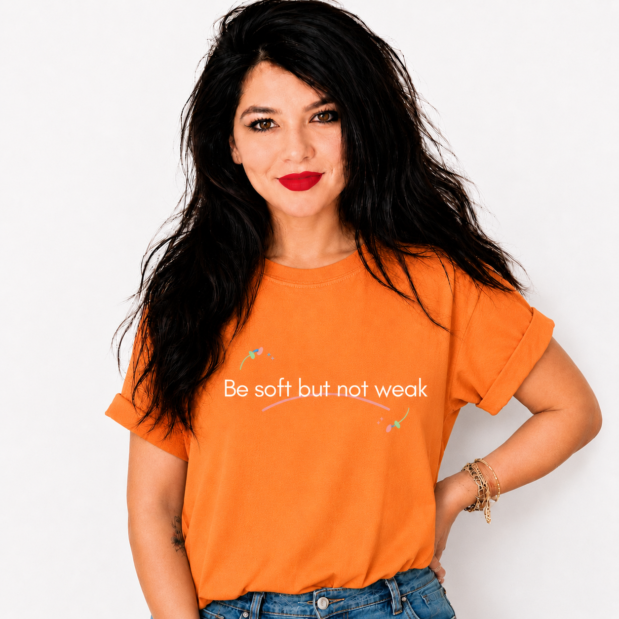 A woman with long black hair wearing a bright orange graphic t-shirt with the text 'Be soft but not weak' printed in white across the chest, paired with blue jeans, against a plain white background.