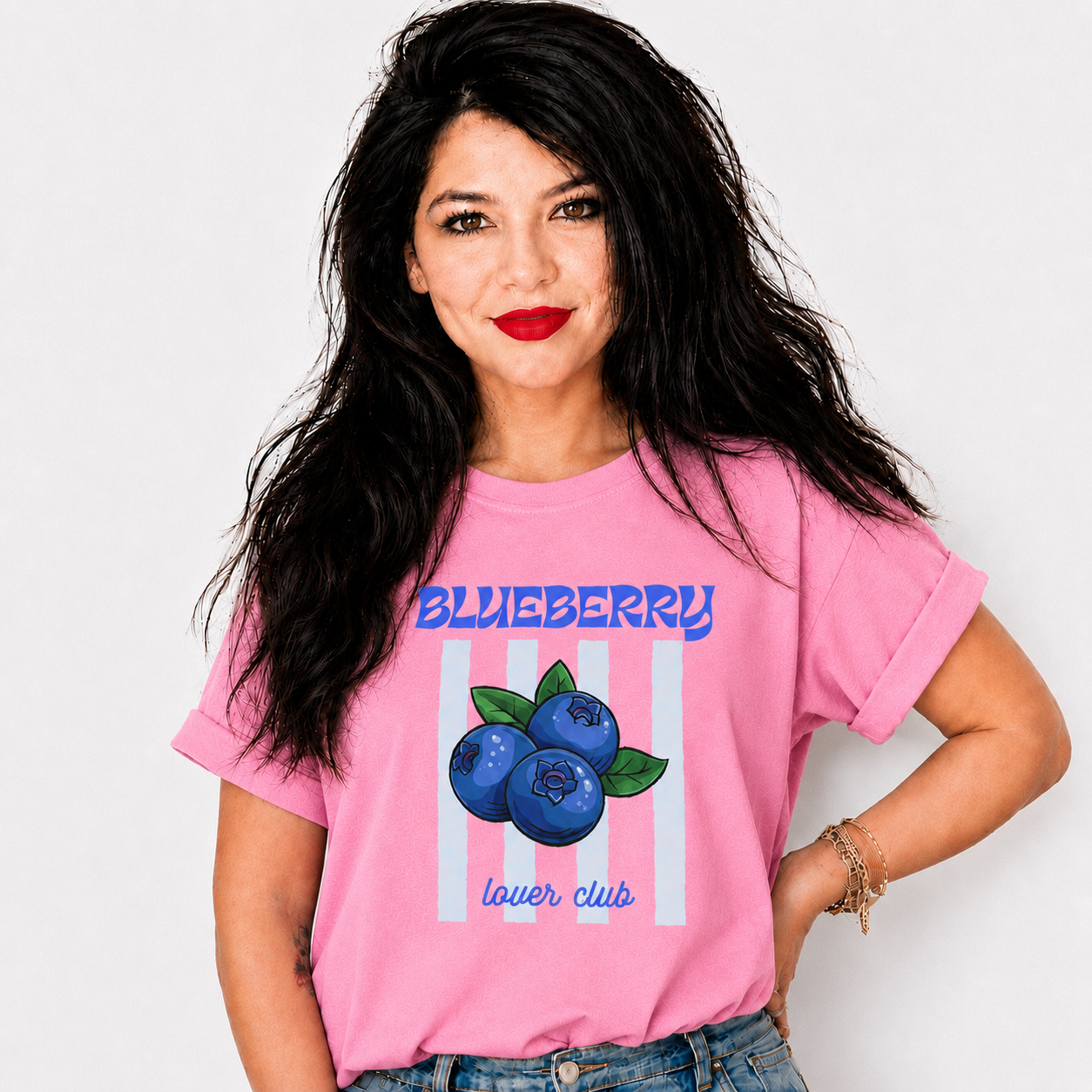 A woman with long black hair wearing a pink graphic t-shirt featuring blueberries and the text 'BLUEBERRY lover club' against a white striped background, paired with blue jeans.