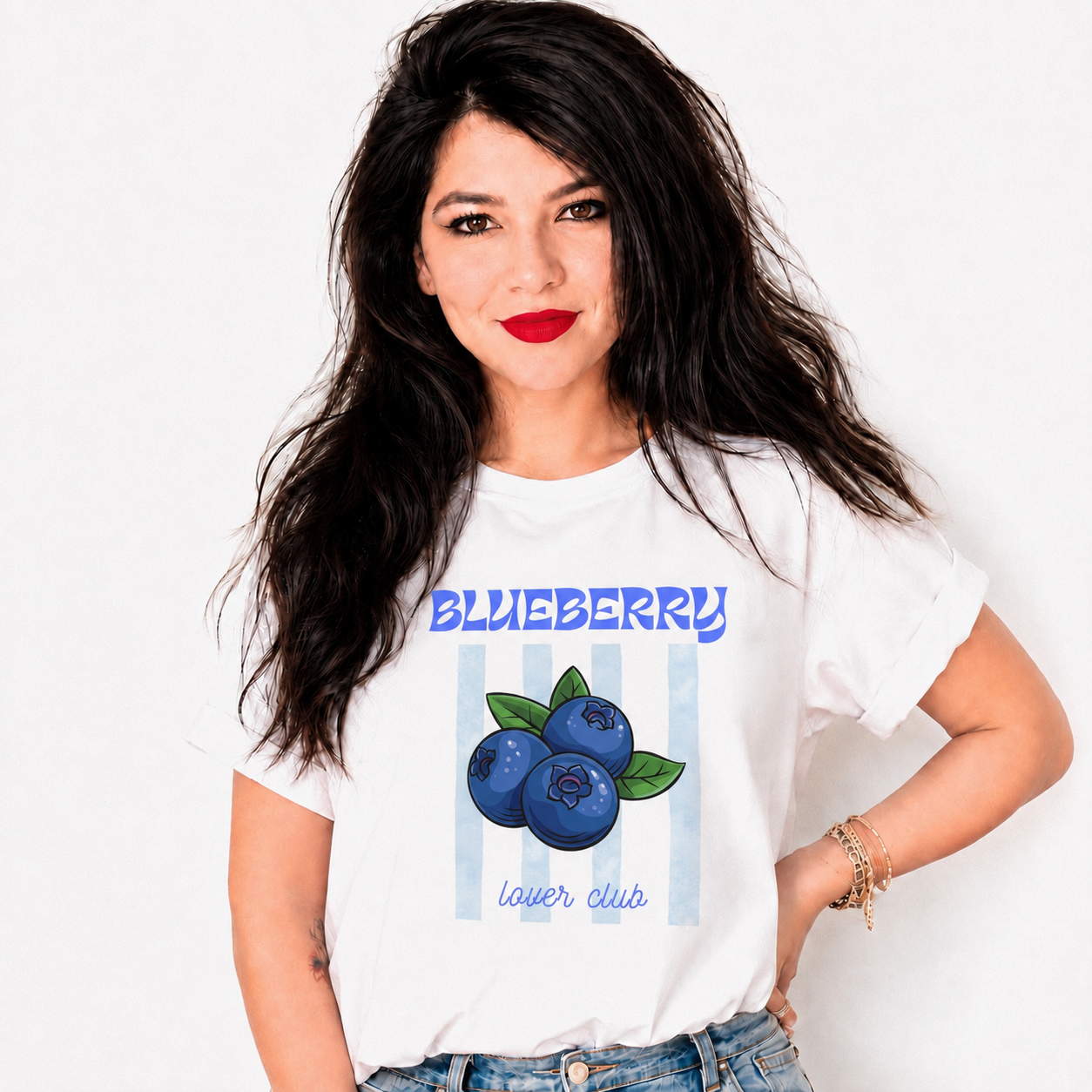 A woman with long dark hair and red lipstick models a white graphic t-shirt featuring a blueberry illustration and the text 'BLUEBERRY lower club' against a light blue striped background. She is wearing blue jeans and gold bracelets, posing against a plain white studio background.