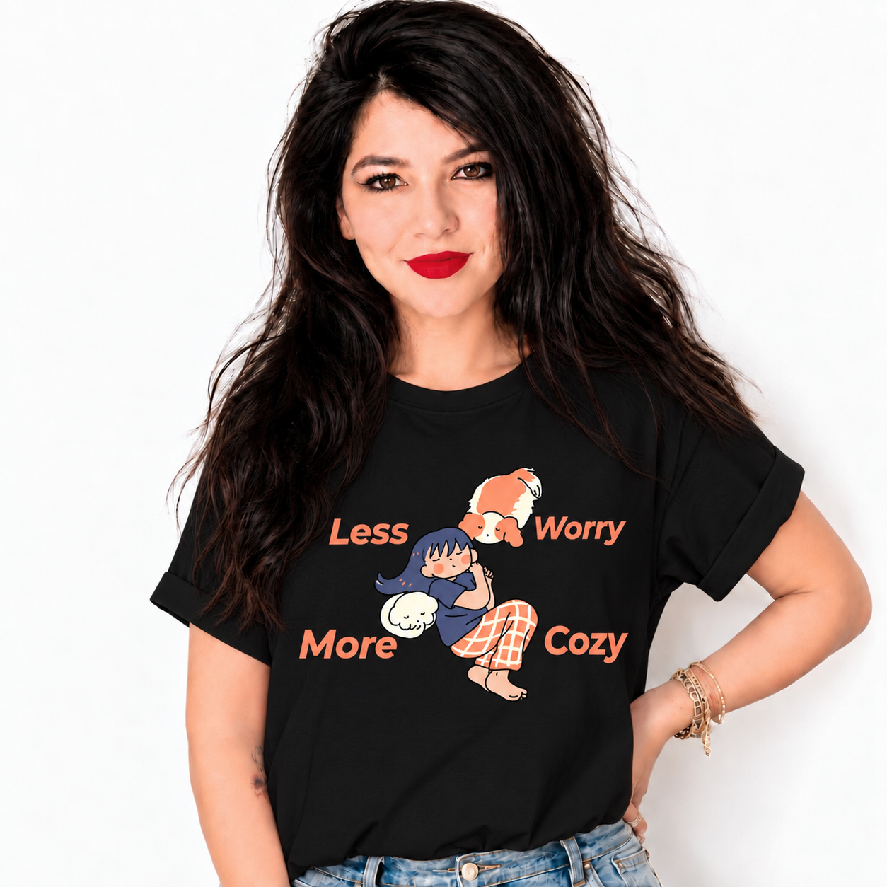 A woman with long dark hair and red lipstick wearing a black graphic t-shirt with the text 'Less Worry More Cozy' and a cartoon illustration of a fox and a girl in pajamas, paired with blue jeans against a plain white background.