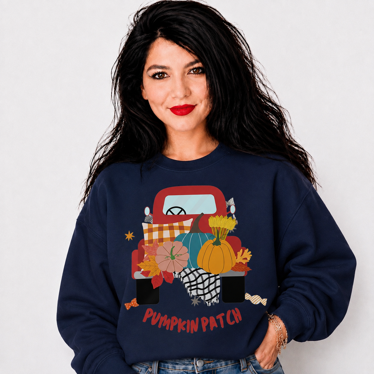 A woman with long black hair and red lipstick models a navy blue crewneck sweatshirt featuring a colorful graphic of a red pickup truck loaded with pumpkins, flowers, and fall foliage, with the text 'PUMPKIN PATCH' printed below.