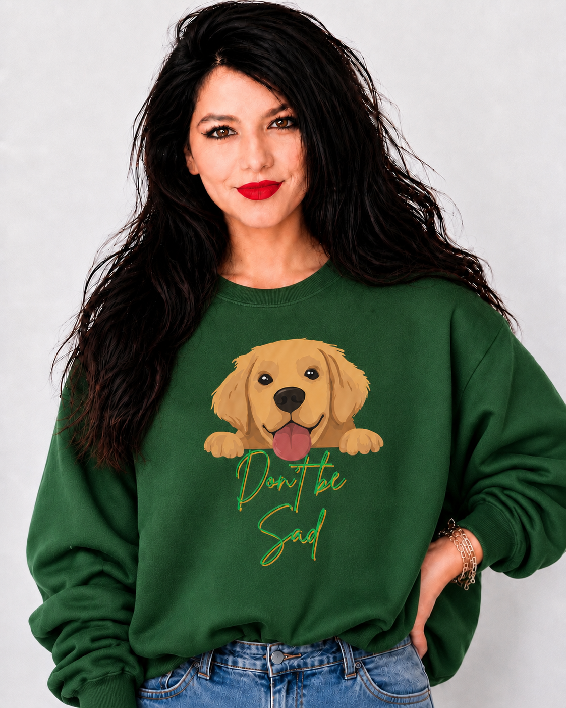 A woman with long dark hair wearing a green crewneck sweatshirt with a cartoon golden retriever graphic and the text 'Don't be sad' in green script, paired with blue jeans against a light gray background.