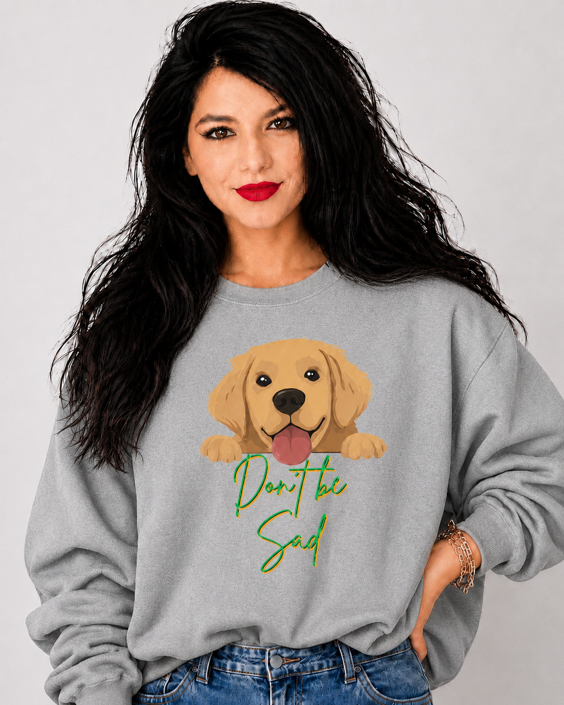 A woman with long black hair wearing a light gray sweatshirt with a cartoon golden retriever graphic and the text 'Don't be sad' in green script, paired with blue jeans against a light gray background.