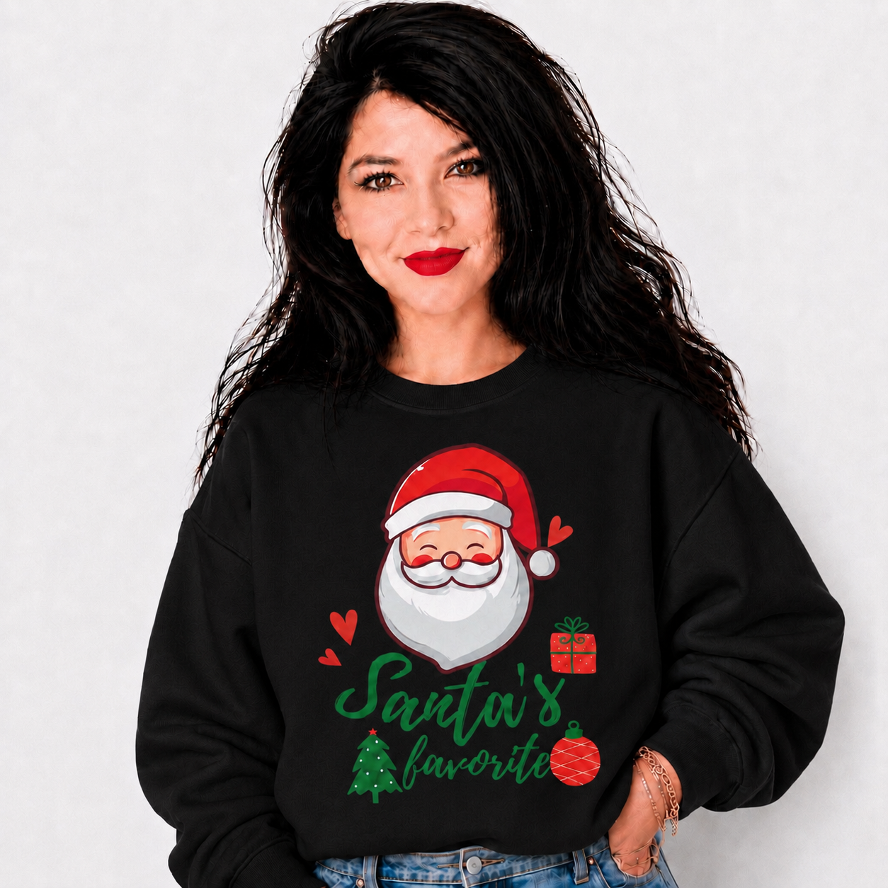 A woman with long black hair wearing a black crewneck sweatshirt featuring a festive graphic of Santa Claus with the text 'Santa's favorite' in green script, accented with red hearts and a small red ornament.