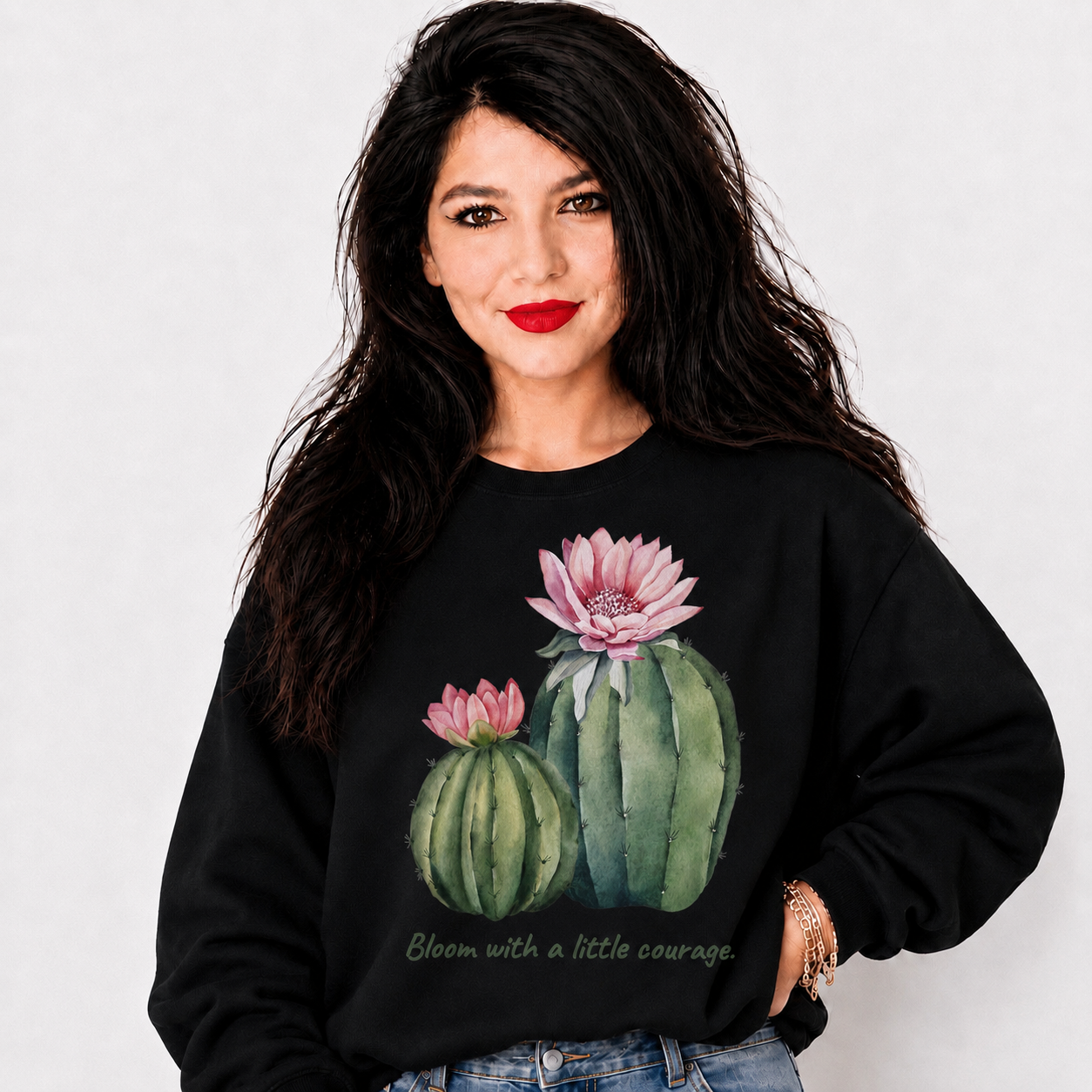 A woman with long dark hair and red lipstick wears a black crewneck sweatshirt featuring a watercolor-style graphic of two green cacti with pink flowers and the text 'Bloom with a little courage' on a light gray background.