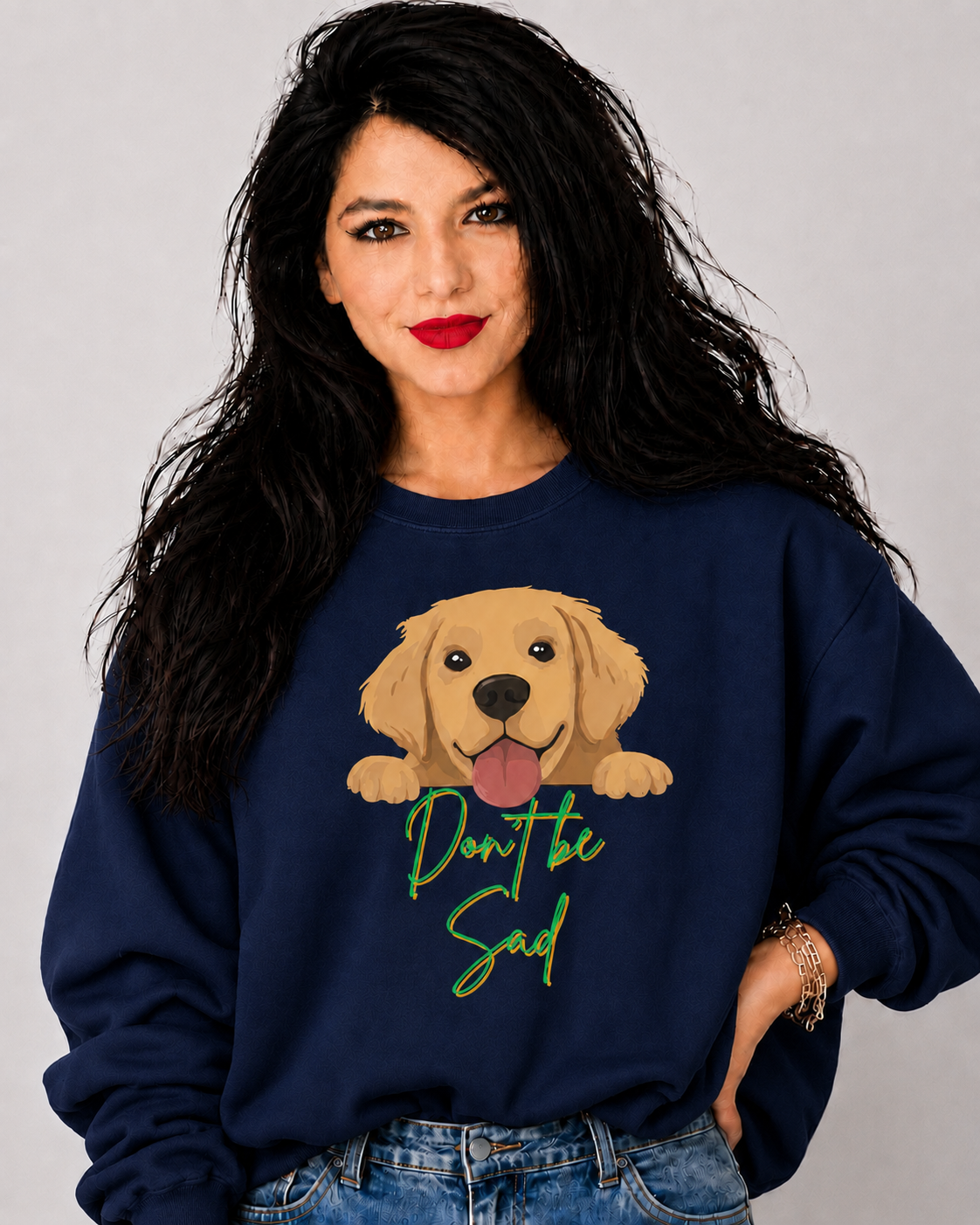 A woman with long black hair wearing a navy blue sweatshirt with a cartoon golden retriever graphic and the text 'Don't be sad' in green script, paired with blue jeans, against a light gray background.