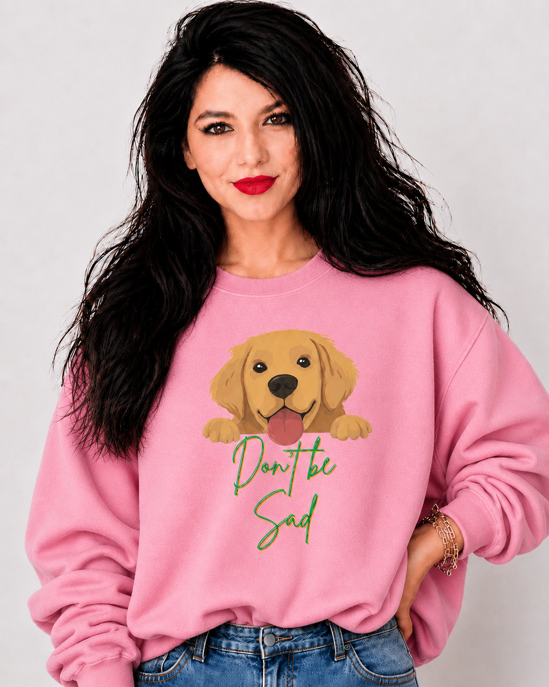 A woman with long black hair wearing a pink crewneck sweatshirt with a cartoon golden retriever graphic and the text 'Don't be sad' in green script, paired with blue jeans against a light gray background.