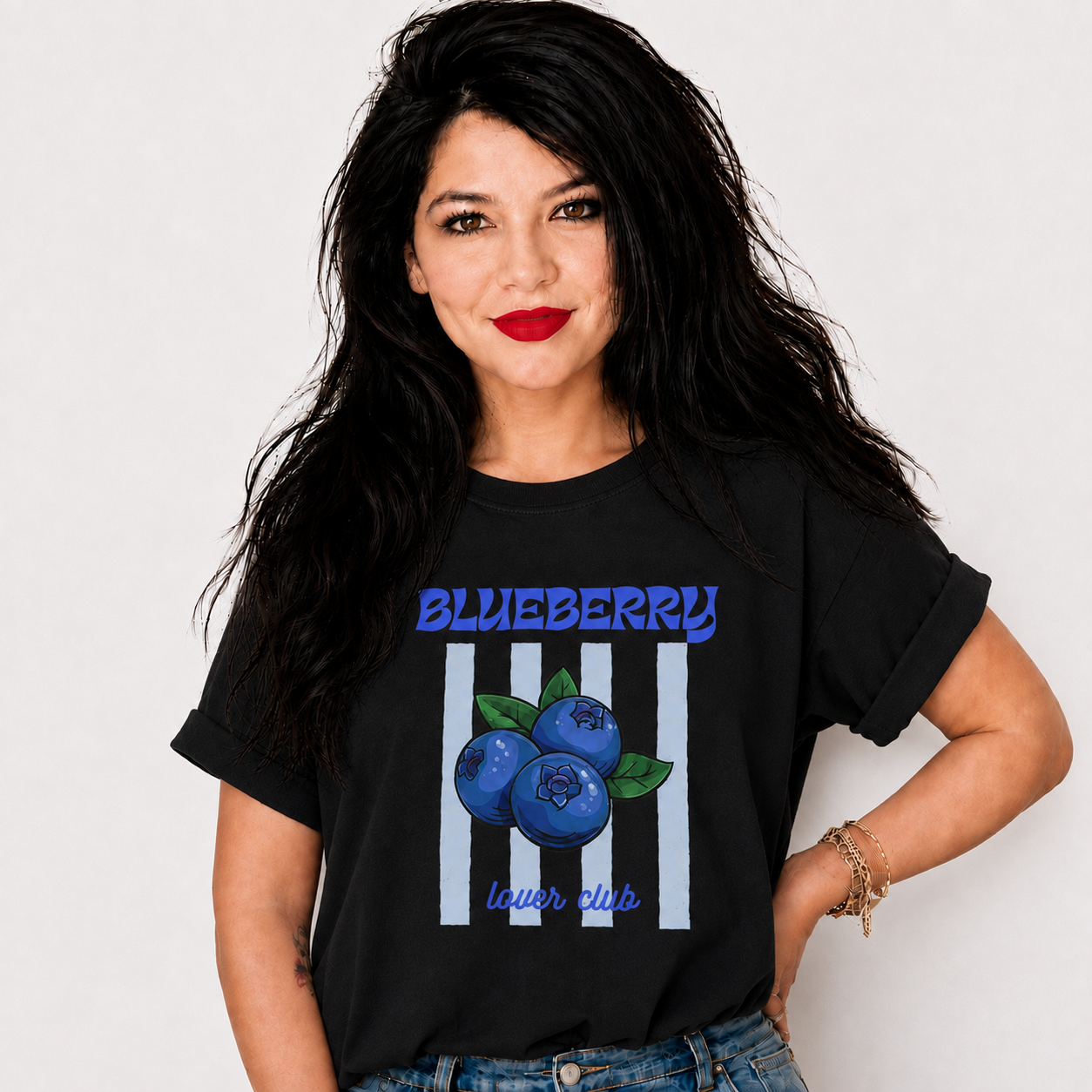 A woman with long black hair and red lipstick wearing a black graphic t-shirt with a blueberry and striped design, paired with blue jeans, against a plain light gray background.