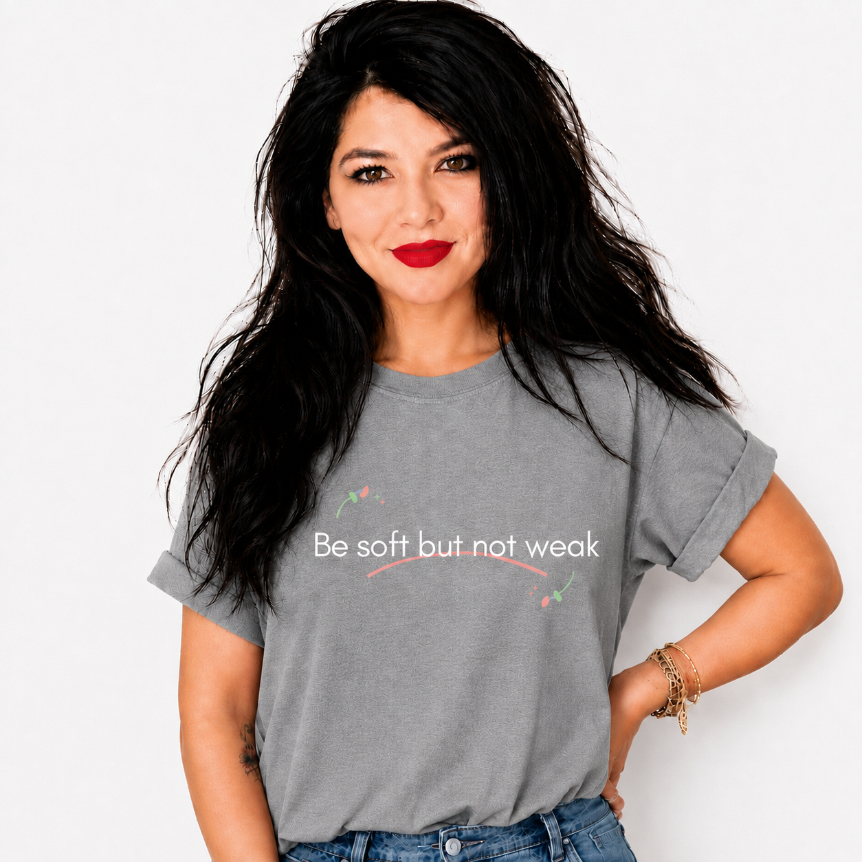 A woman with long black hair and red lipstick wearing a gray graphic t-shirt with the text 'Be soft but not weak' printed in white, paired with blue jeans, against a plain white background.