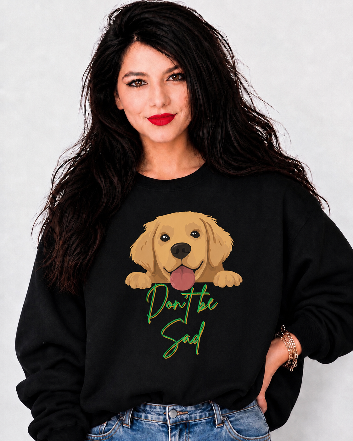 A woman with long dark hair wearing a black crewneck sweatshirt featuring a cartoon golden retriever graphic and the text 'Don't be Sad' in green script, paired with blue jeans against a light gray background.