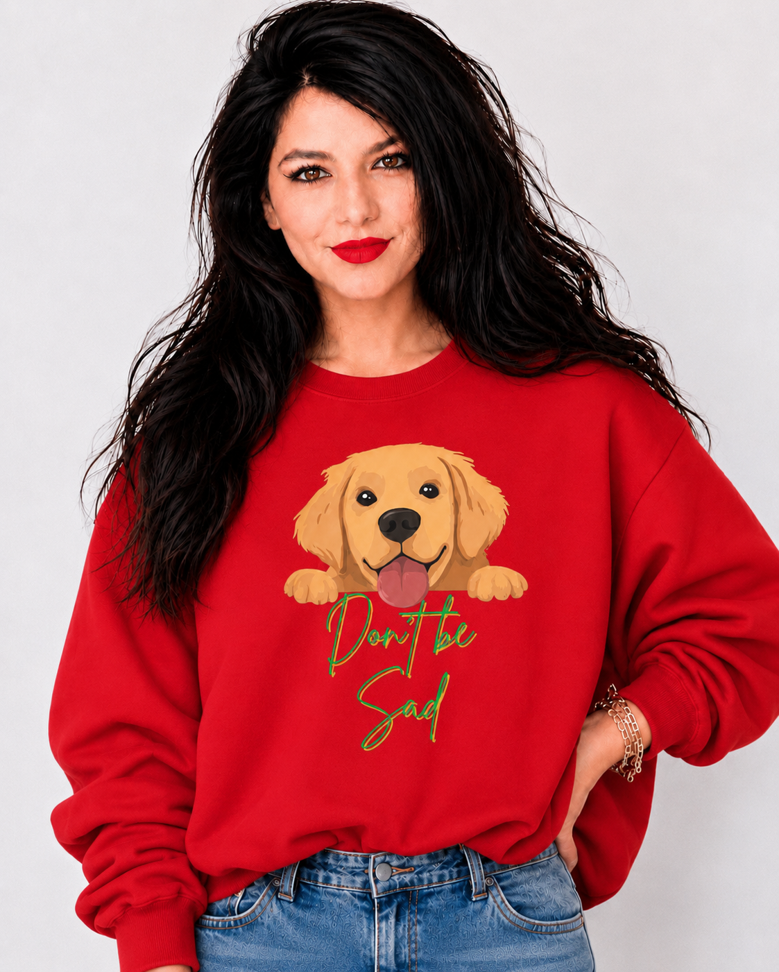A woman with long black hair wearing a red crewneck sweatshirt with a golden retriever graphic and the text 'Don't be sad' in green, paired with blue jeans, against a light gray background.