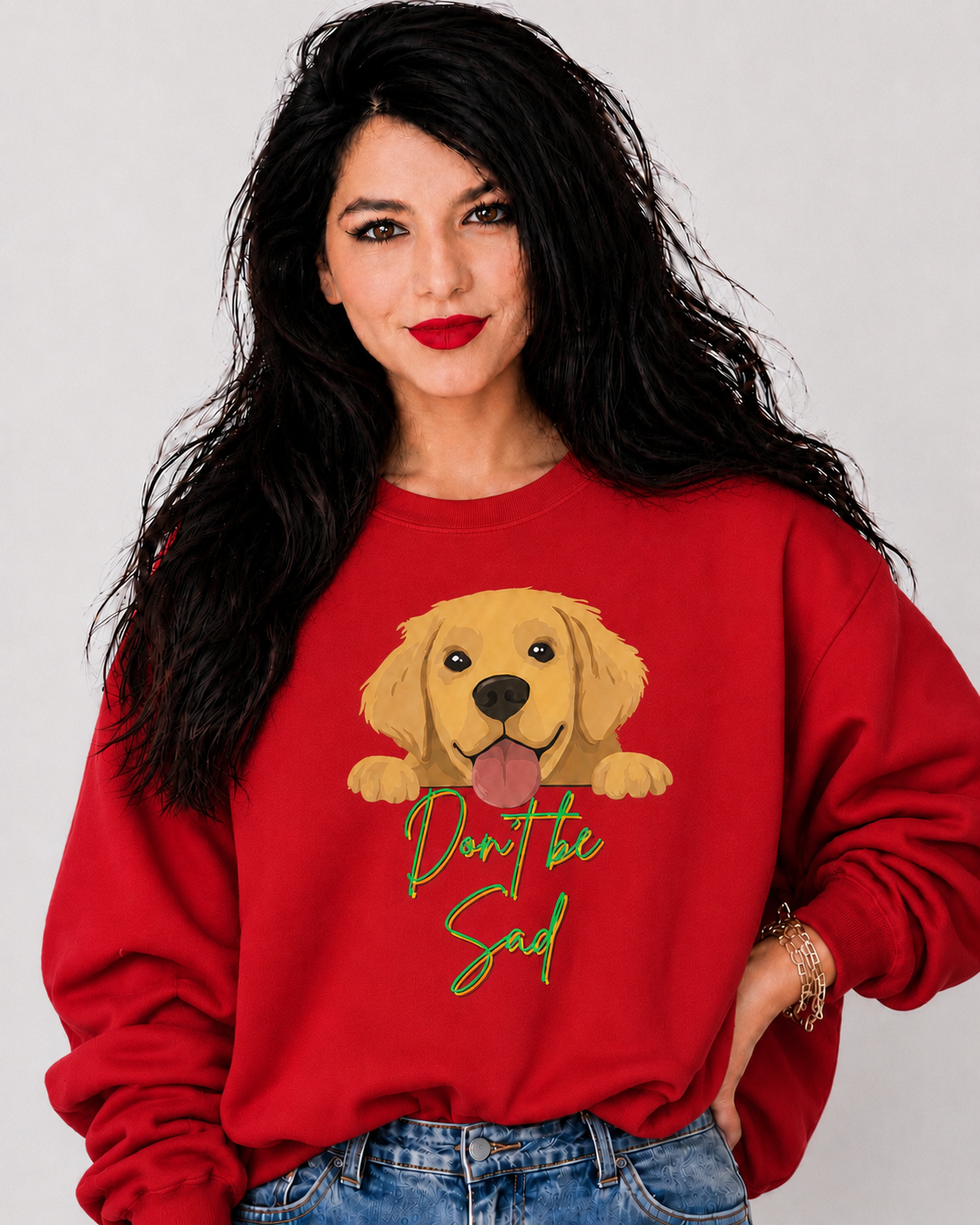 A woman with long black hair wearing a red crewneck sweatshirt featuring a cartoon golden retriever graphic and the text 'Don't be sad' in green script, paired with blue jeans against a light gray background.