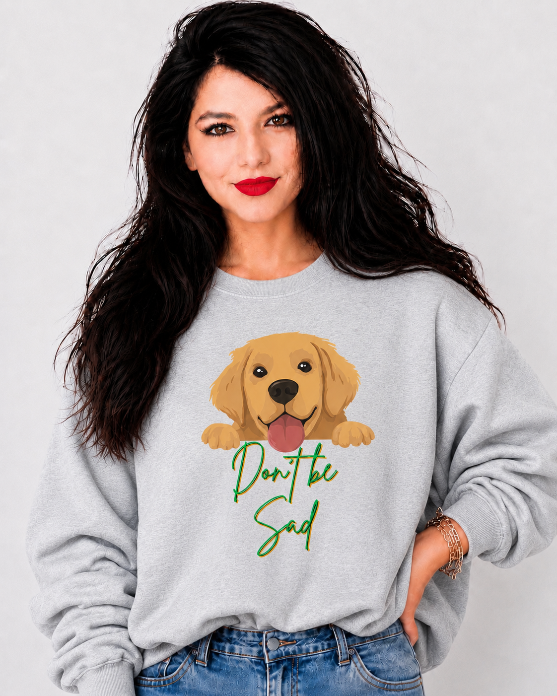 A woman with long dark hair wearing a light gray crewneck sweatshirt with a cartoon golden retriever graphic and the text 'Don't be sad' in green script, paired with blue jeans against a light gray background.