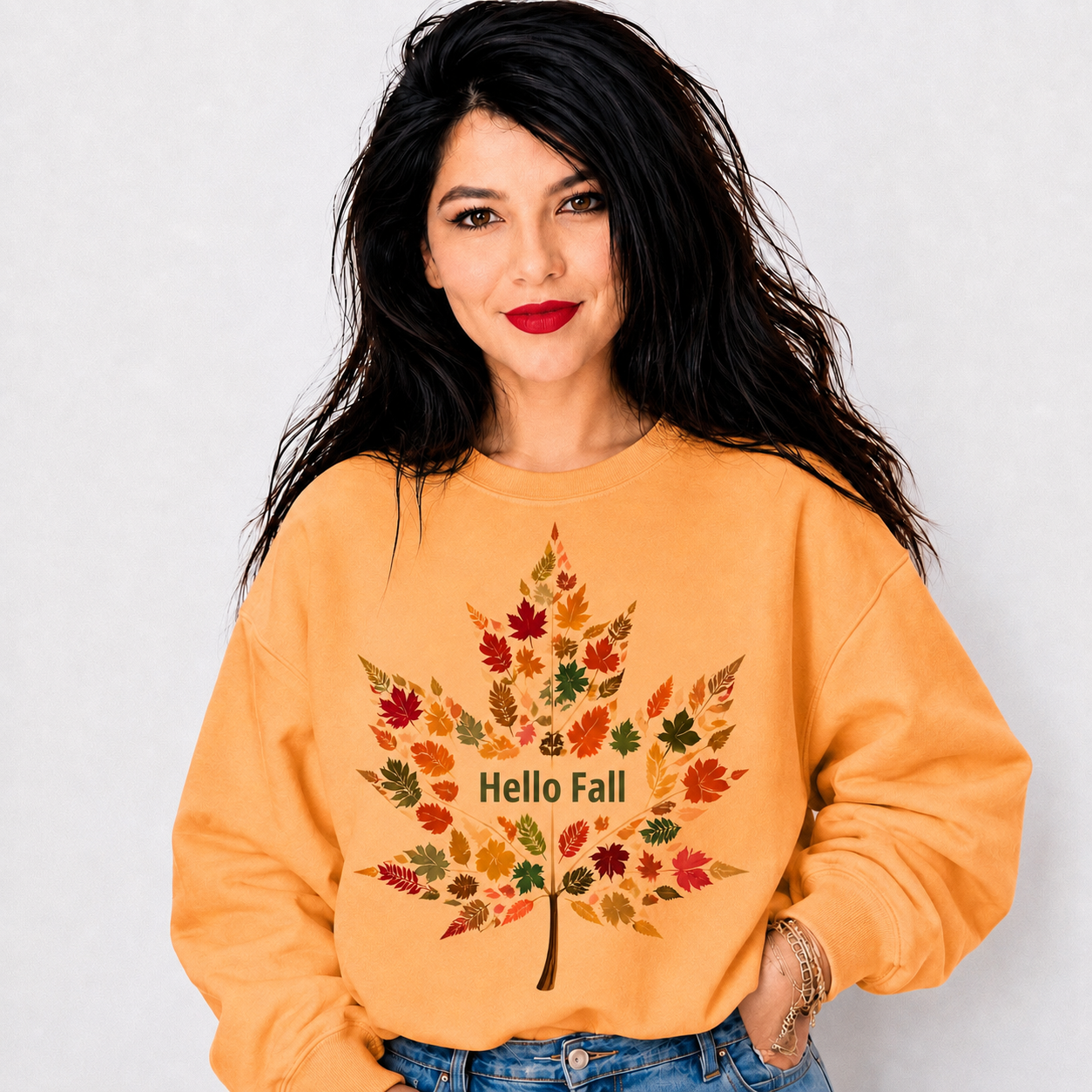 A woman with long black hair wearing a mustard yellow sweatshirt with a colorful autumn leaf graphic and the text 'Hello Fall' on the front, paired with blue jeans against a light gray background.