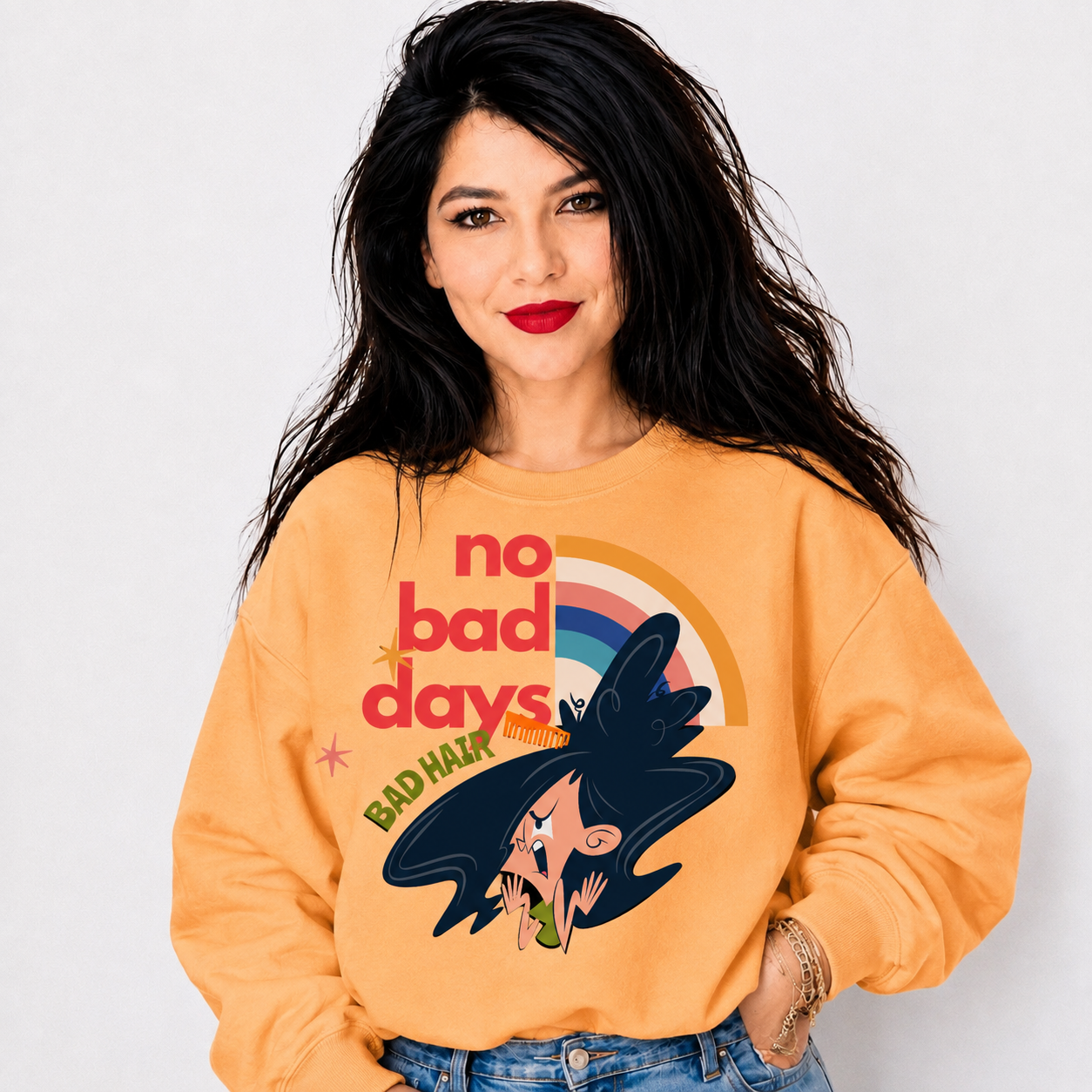 A woman with long black hair wearing a light orange sweatshirt with a graphic that reads 'no bad days' and 'BAD HAIR' alongside a cartoon witch with dark hair and a rainbow. She is wearing blue jeans against a plain light gray background.