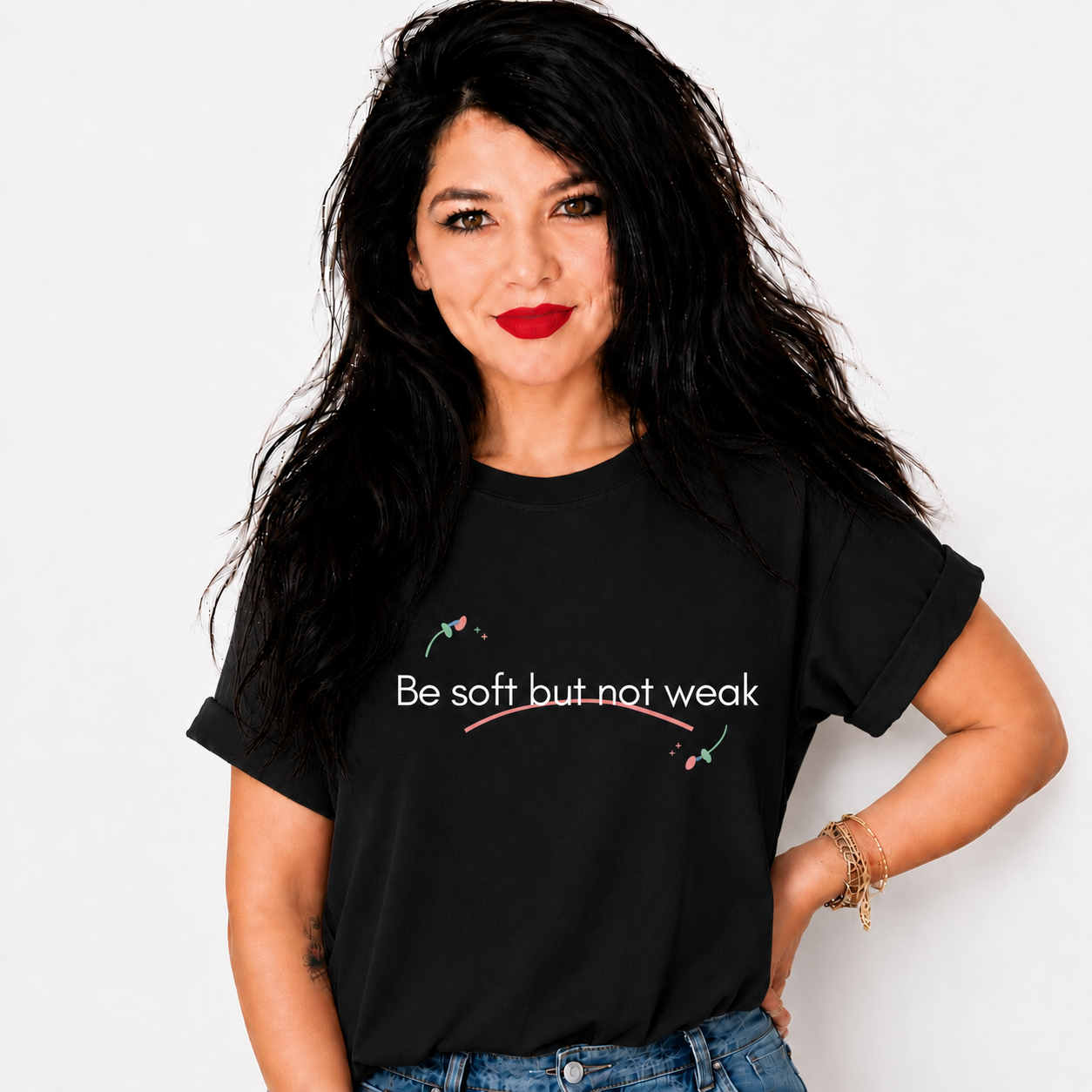 A woman with long, dark, messy hair is wearing a black graphic t-shirt with the text 'Be soft but not weak' printed in white, accented by small pink and green floral details. She is posing against a clean, off-white background.