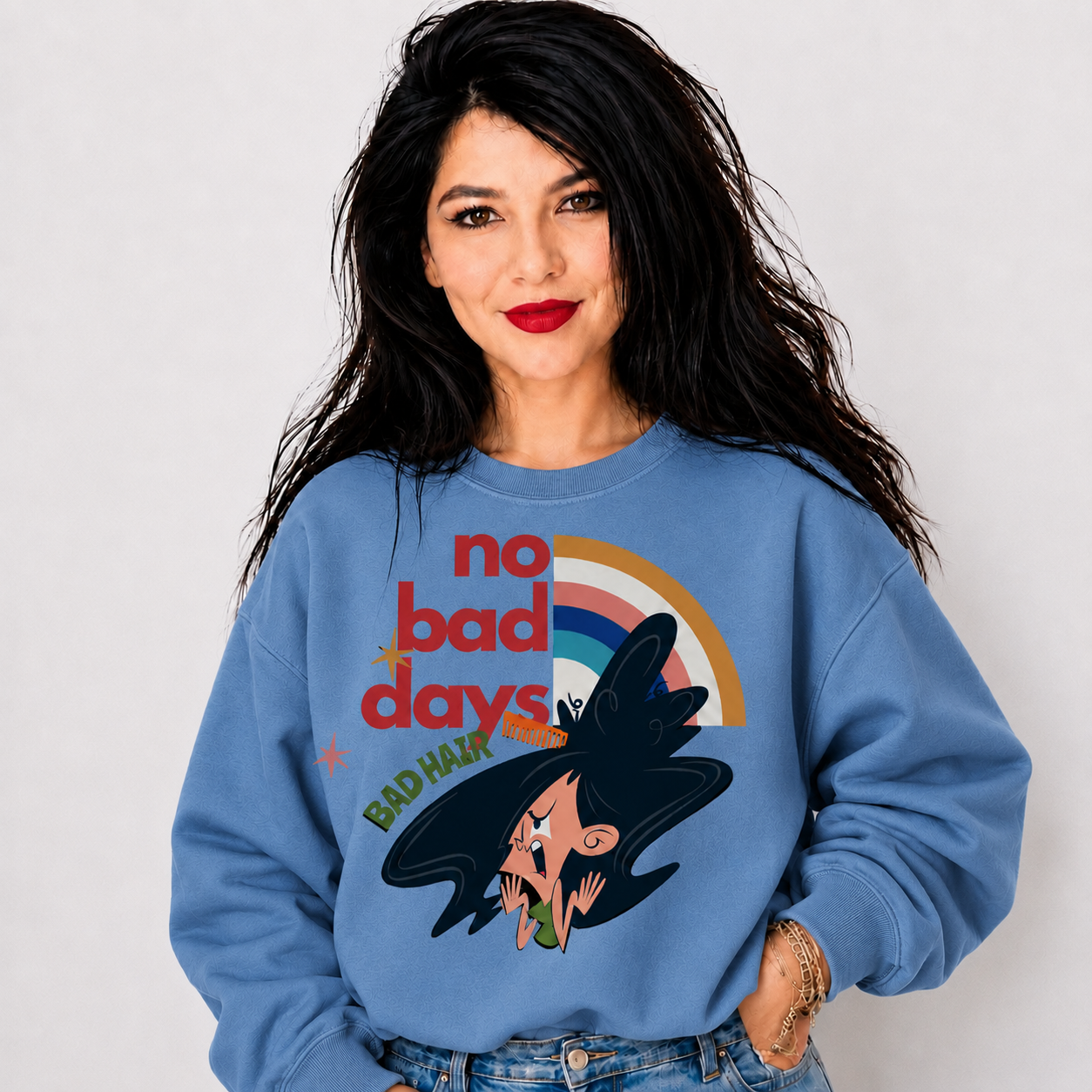 A woman with long black hair wearing a light blue crewneck sweatshirt with a retro graphic that reads 'no bad days' and 'BAD HAIR' alongside a cartoon witch with a rainbow. She is posing against a light gray studio background.