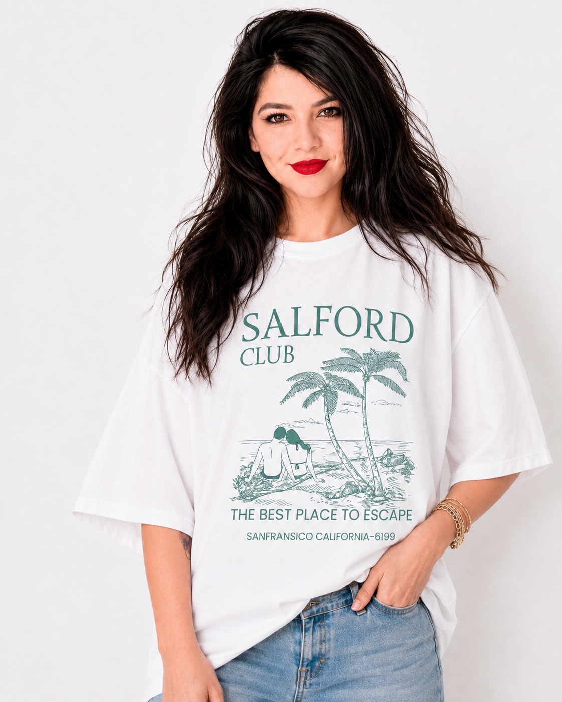 A woman wearing a white oversized graphic t-shirt with 'SALFORD CLUB' printed in green on the front, paired with light blue jeans, against a plain white background.