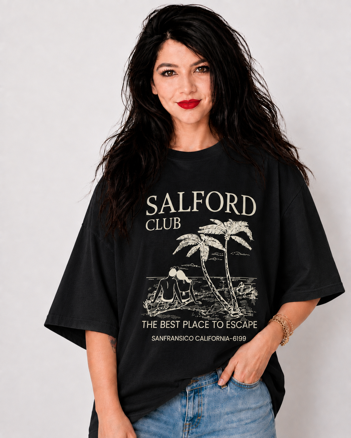 A woman with long dark hair wearing a black oversized graphic t-shirt with 'SALFORD CLUB' printed in white on the front, paired with blue jeans, against a light gray background.