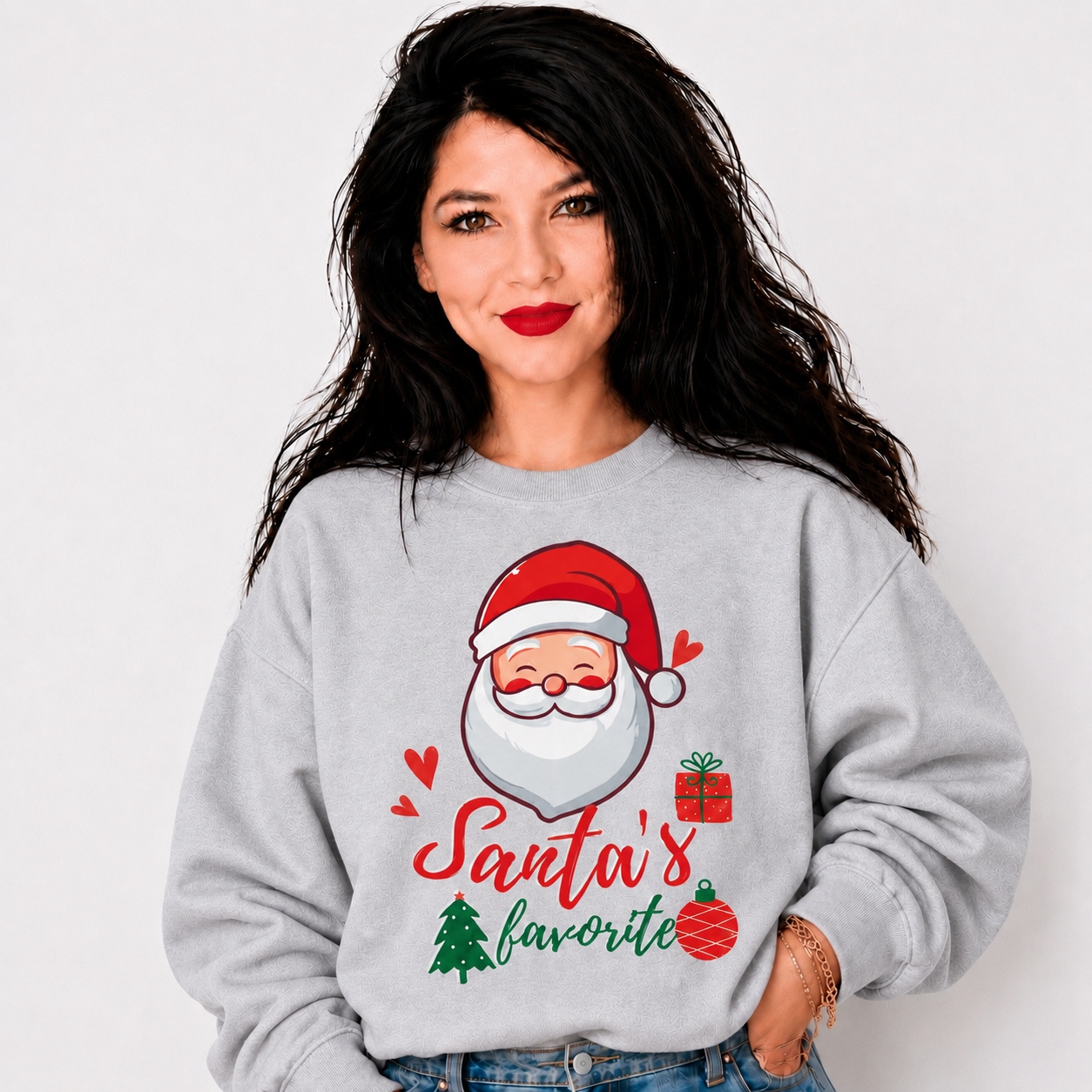 A woman wearing a light gray crewneck sweatshirt with a festive graphic of Santa Claus and the text 'Santa's favorite' in red and green, paired with blue jeans against a plain white background.