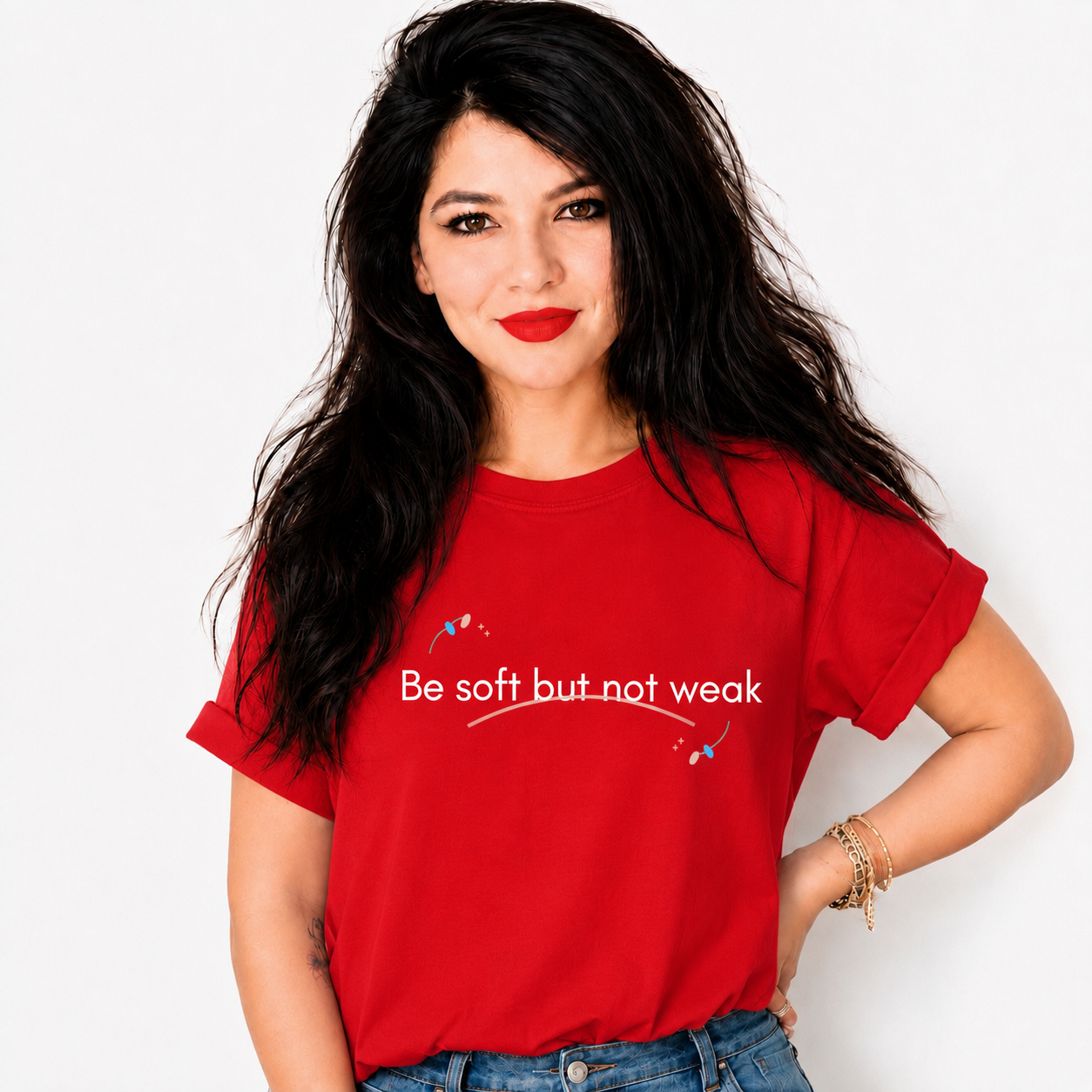 A woman with long black hair and red lipstick wearing a red graphic t-shirt with the text 'Be soft but not weak' printed in white across the chest, paired with blue jeans against a plain white background.