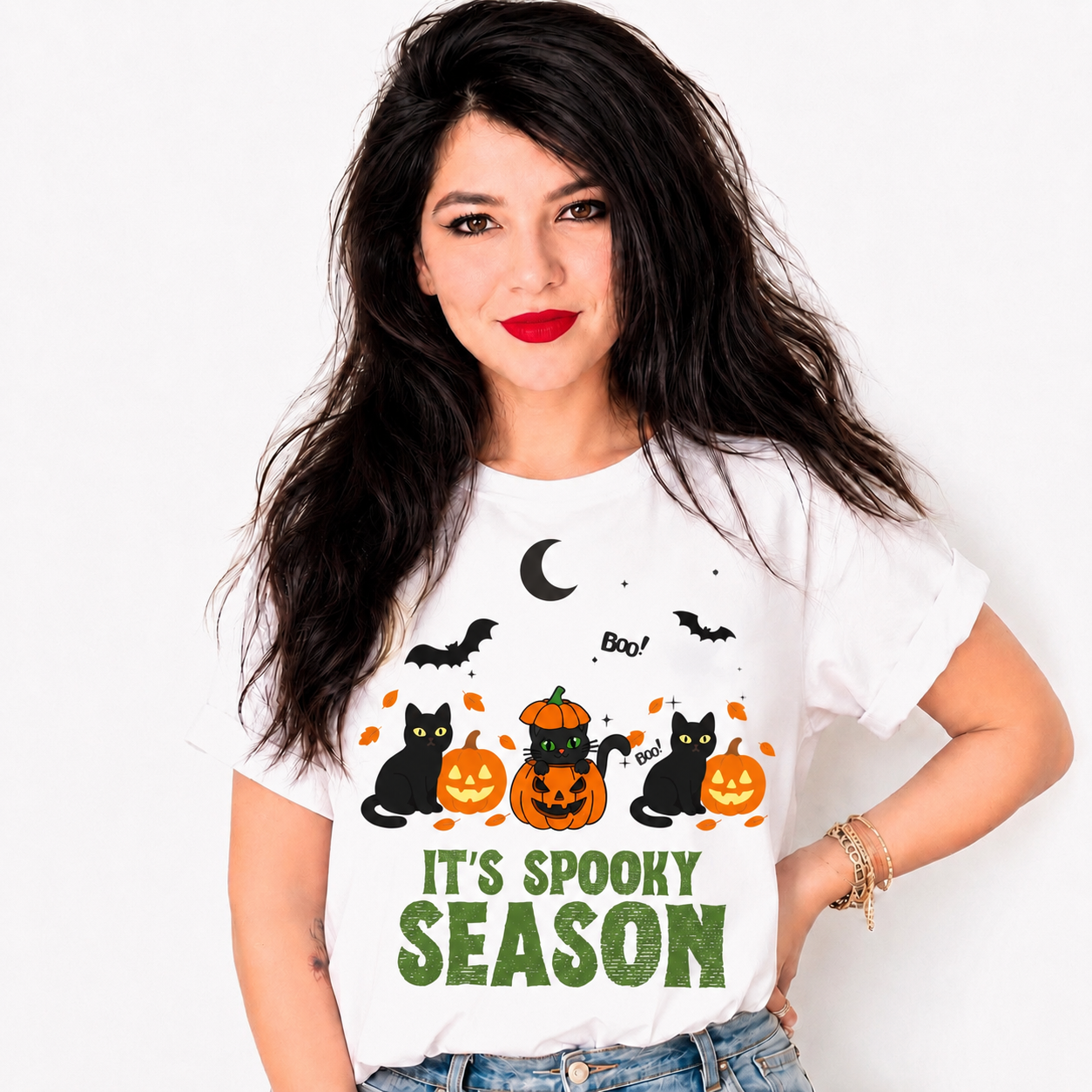 A woman with long dark hair wearing a white graphic t-shirt with a Halloween design. The design features black cats, jack-o'-lanterns, bats, a crescent moon, and the text 'IT'S Spooky SEASON' in green.