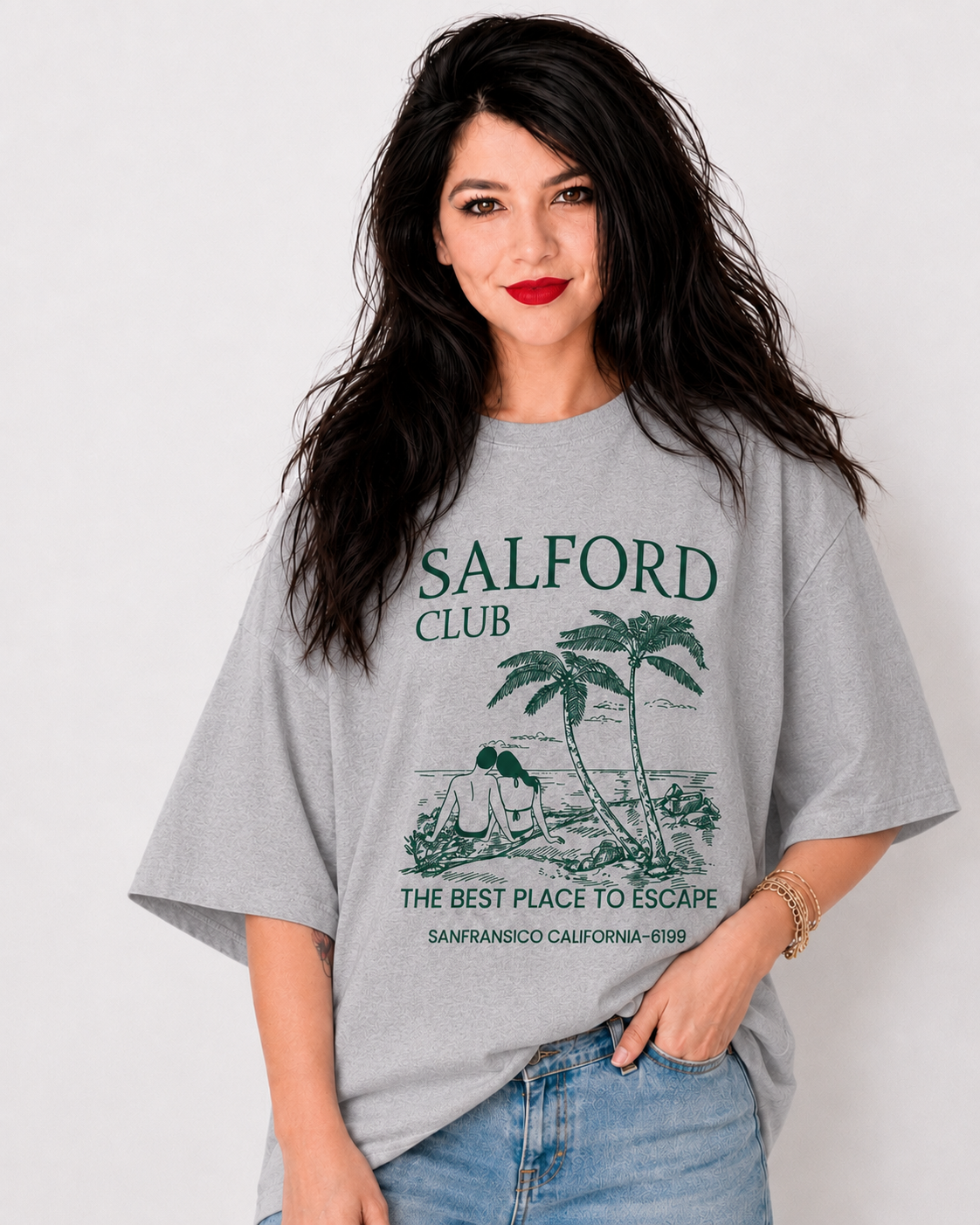 A woman wearing an oversized light gray graphic t-shirt with 'SALFORD CLUB' and a palm tree illustration printed in dark green, paired with light blue jeans against a plain white background.