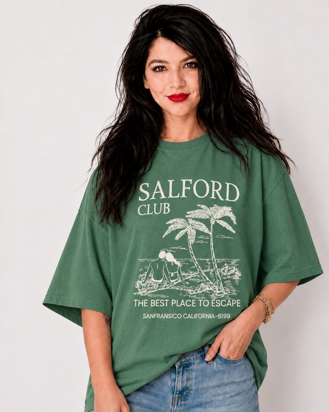 A woman with long dark hair wearing an oversized sage green graphic t-shirt with 'SALFORD CLUB' printed in white on the front, paired with blue jeans, against a plain light gray background.