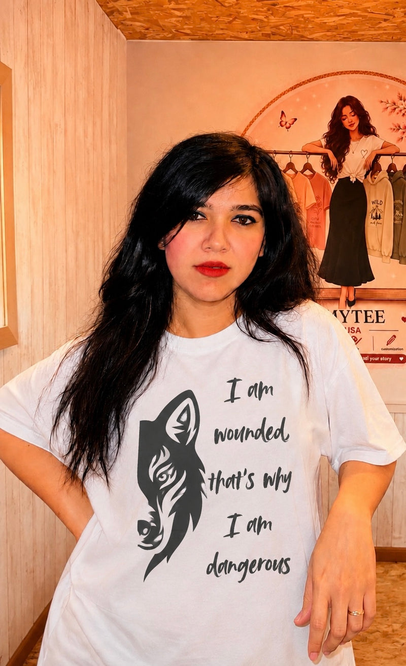 A woman with long black hair wearing a white graphic t-shirt with a black wolf illustration and the text 'I am wounded that's why I am dangerous'. She is standing in a room with light-colored wooden walls and a poster in the background.